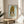 May include: Framed poster featuring anime-style illustration of young girl with large golden blonde hair in tall beehive style, oversized eyes, and yellow top against pale blue background. Yoshitomo Nara exhibition text below artwork. Natural wood frame sits on round wooden side table next to beige upholstered chair with plant leaves visible, warm sunlight streaming across neutral-toned room.