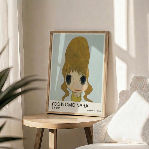May include: Framed poster featuring anime-style illustration of young girl with large golden blonde hair in tall beehive style, oversized eyes, and yellow top against pale blue background. Yoshitomo Nara exhibition text below artwork. Natural wood frame sits on round wooden side table next to beige upholstered chair with plant leaves visible, warm sunlight streaming across neutral-toned room.