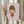 May include: Hands holding a framed art print featuring a minimalist illustration of a young girl with brown bob haircut, closed eyes, and red dress with her tongue sticking out, set against a soft pink background. The artwork is displayed in a simple wooden frame. Person wearing cream-colored ruffled sleeves holds dried wheat stalks in one hand while positioning the framed print against white curtains in soft natural lighting.