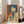May include: Large framed poster leaning against white wall next to beige sofa in bright living room with hardwood floors and large windows. The vintage-style artwork features a woman with dark bob haircut and red lips holding a handgun, rendered in orange, blue and cream colors with Japanese text, displayed in white frame.