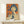 May include: Framed vintage-style Japanese Coca-Cola advertisement poster featuring a woman with dark bob haircut holding a Coke bottle against orange background with blue Japanese text, displayed on light wood console table next to small white wrapped gift with pine sprig, against cream wall with natural lighting