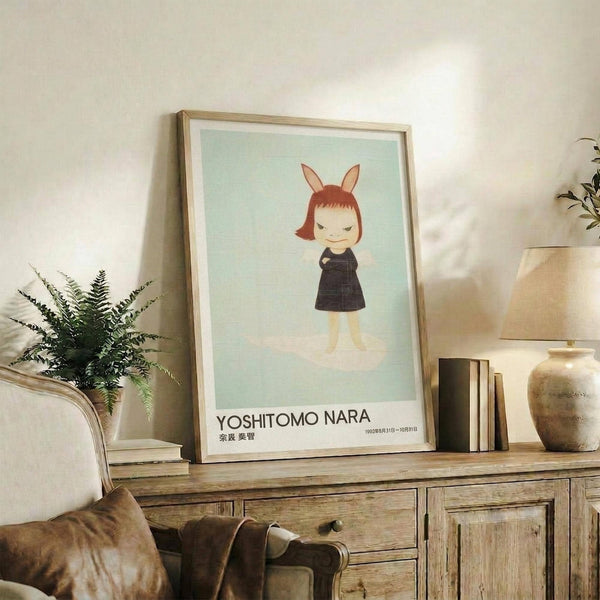 May include: Framed poster displaying a stylized illustration of a small figure with reddish-brown hair and bunny ears wearing a dark dress on pale mint green background, positioned on rustic wooden console table beside fern plant, ceramic vase, and stacked books in warm-toned living room setting with beige armchair