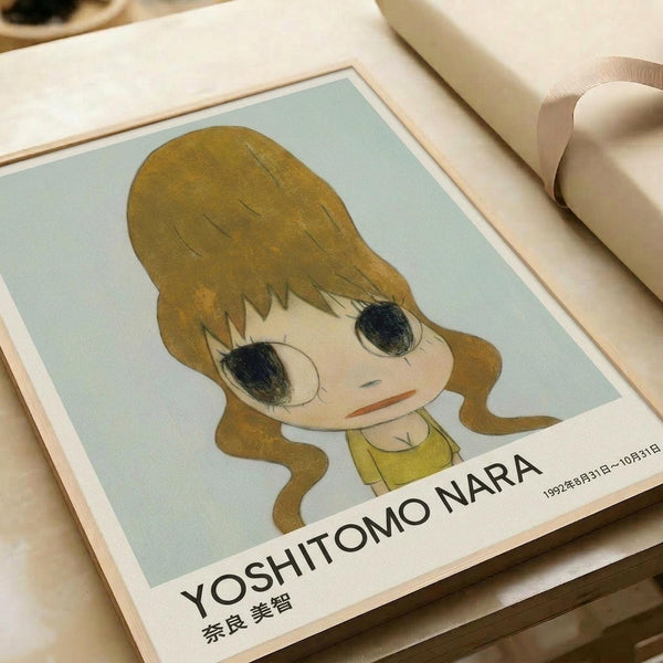 May include: Framed art print featuring a cartoon-style illustration of a young girl with large dark eyes and golden brown hair against a pale mint green background. The print shows "YOSHITOMO NARA" text at bottom with Japanese characters and dates. Frame appears light wood-toned, displayed on cream colored bedding or fabric surface with soft natural lighting.