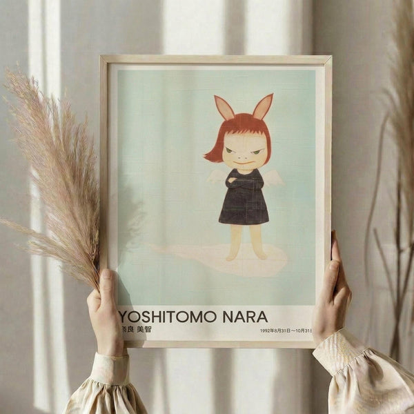 May include: Hands holding a framed art print featuring a cartoon child character with reddish-brown bob haircut and bunny ears, wearing a dark dress against a soft mint green background. The print shows "YOSHITOMO NARA" text and Japanese characters below the artwork. Person also holds dried wheat or pampas grass stems. Light wooden frame with white matting, photographed against neutral beige curtains in soft natural lighting.