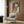 May include: Framed vintage-style poster featuring a woman with blue bob haircut holding a gun, rendered in orange and blue pop art aesthetic with Japanese text. Displayed in modern living room setting above beige armchair with geometric pillow, alongside brass floor lamp and dark side table with open book.