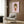 May include: A framed poster featuring a cartoon-style illustration of a young girl with brown bob hair and closed eyes, wearing a red dress, sticking out her tongue on a soft pink background. The artwork is displayed on a wood-paneled wall above a beige upholstered armchair with geometric patterned pillow, beside a tall brass floor lamp and dark round side table with an open book.