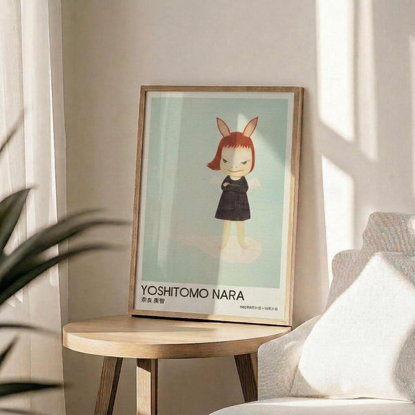 May include: Framed art print featuring a stylized character with reddish bob haircut, bunny ears, and dark dress against pale green background, displayed on round wooden side table next to beige upholstered chair with white pillow, green plant leaves visible in corner, natural sunlight casting shadows on neutral wall