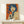 May include: Framed vintage-style poster with orange background featuring woman with blue bob haircut holding gun, displayed on light wood shelf next to small white gift box with greenery sprig against cream wall