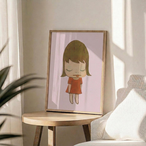 May include: Framed art print displayed on wooden round side table next to beige upholstered chair with white pillow. Print shows cartoon-style illustration of young girl with brown bob haircut, closed eyes, and red dress on soft pink background. Natural lighting creates window shadows on neutral wall behind. Green plant leaves visible in corner.