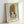 May include: Framed art print featuring a cartoon-style illustration of a young girl with golden blonde hair in an updo and side curls, large dark eyes, and yellow top against a pale mint green background. The print shows "YOSHITOMO NARA" text at bottom with Japanese characters and dates. Natural wood frame displayed leaning against white wall, showing subtle shadows and lighting.