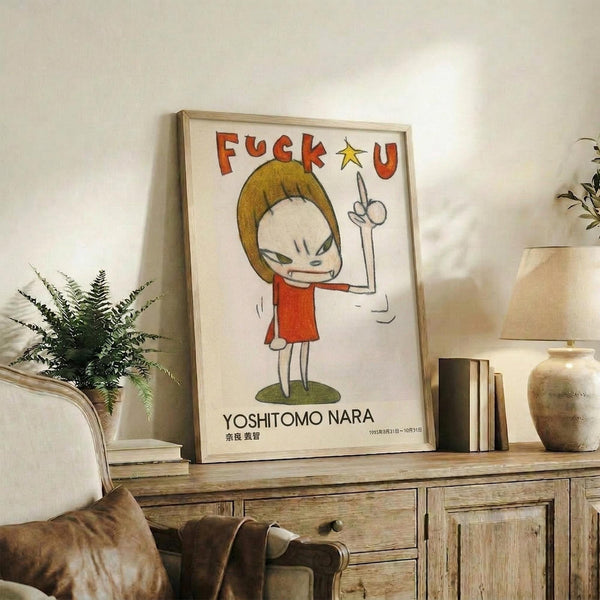 May include: Framed poster featuring a cartoon character with blonde hair wearing a red dress, giving the middle finger with "F*CK U" text above and "YOSHITOMO NARA" below. The artwork is displayed on a rustic wooden sideboard next to a fern plant, pottery vase, and books in a cozy living room setting with neutral tones and natural lighting.