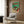 May include: Vintage Japanese turntable poster with green and red retro design featuring record player illustration, displayed in light wood frame on brown wood panel wall above beige upholstered armchair with geometric patterned pillow, modern brass floor lamp and side table with open book nearby