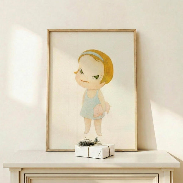 May include: Framed art print featuring a stylized cartoon child with blonde bob haircut and green eyes, wearing a light blue dress while holding a small pig. The illustration has soft, muted colors on cream background. Displayed in thin wooden frame on white wooden console table alongside small wrapped gift with evergreen sprig. Neutral wall backdrop with natural lighting.