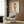 May include: Framed poster featuring cartoon-style character with blonde hair in red dress making peace sign, with colorful text above and artist name below. Shown in modern living room setting with beige upholstered chair, geometric patterned pillow, brass floor lamp, and round black side table with open book against wood-paneled wall.