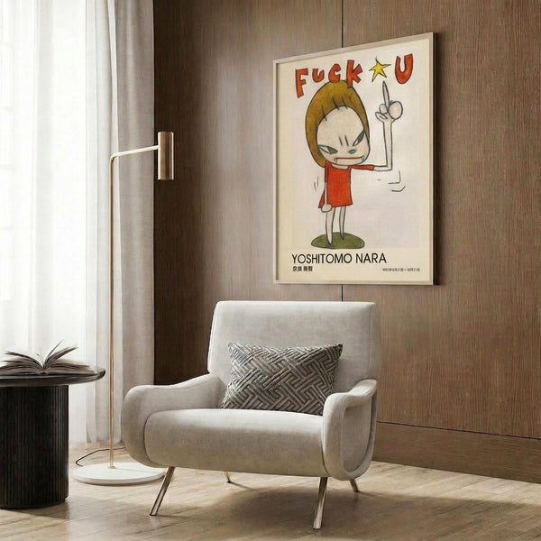 May include: Framed poster featuring cartoon-style character with blonde hair in red dress making peace sign, with colorful text above and artist name below. Shown in modern living room setting with beige upholstered chair, geometric patterned pillow, brass floor lamp, and round black side table with open book against wood-paneled wall.