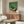 May include: Framed vintage Japanese turntable advertisement poster with green and red design hanging above mid-century walnut credenza in minimalist living room. White upholstered chair and glass-top coffee table visible. Poster features retro record player illustration with Japanese text against vibrant green background.