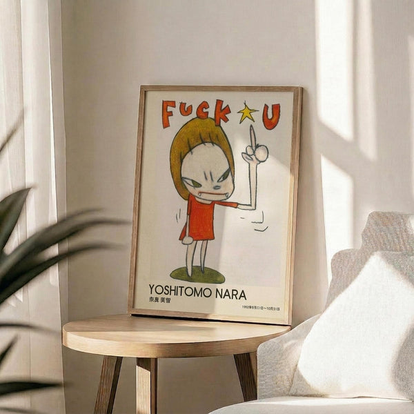 May include: Framed art print displayed on round wooden side table next to beige upholstered chair and potted plant. The poster shows a cartoon-style character with blonde hair wearing red shirt, making an angry expression with raised middle finger. Text reads "FUCK U" at top and "YOSHITOMO NARA" at bottom. Print has thin wooden frame and white matting, positioned in bright room with natural lighting and neutral decor.