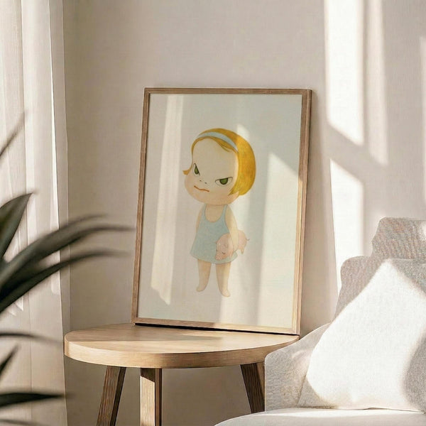 May include: Framed artwork showing a stylized cartoon character with blonde bob haircut, green eyes, and light blue dress, displayed on a round wooden side table next to a beige upholstered chair. Natural sunlight streams across the scene, casting soft shadows on white walls. A plant's leaves are visible in the foreground, creating an airy, minimalist home decor setting.