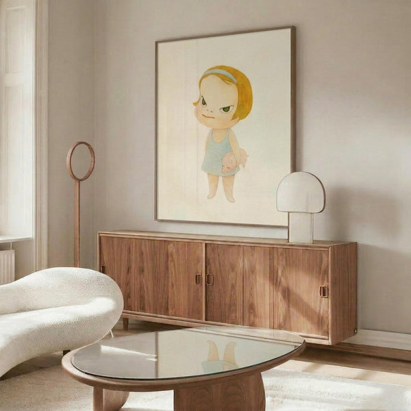 May include: Framed art print displayed on white wall above mid-century walnut credenza in modern living room. Print features cartoon-style character with blonde bob haircut and light blue outfit on cream background. Room has white curved chair, glass coffee table, and minimalist decor with natural lighting.