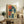 May include: Framed vintage-style poster featuring a woman with tousled dark hair drinking from a cup, rendered in cream, teal and red colors with Japanese characters. Shown leaning against white wall on rustic wooden sideboard alongside potted fern, books, and ceramic vases in neutral-toned living room setting.