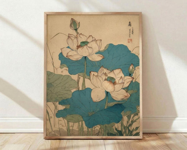 May include: Framed vintage Japanese botanical print featuring two cream-colored lotus flowers with teal centers surrounded by large blue-green lotus leaves. The artwork shows delicate line work and muted colors on aged paper background with Japanese characters in upper right corner. Natural wood frame sits on light wooden floor against white wall with soft shadows.