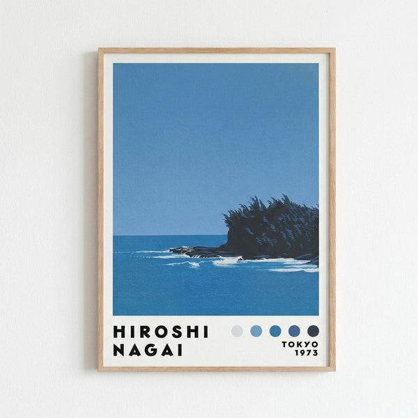 May include: Framed art print hanging on white wall showing coastal scene with dark rocky outcrop and vegetation against blue ocean and sky, featuring bold graphic style in blues and teals with white matting and light wood frame