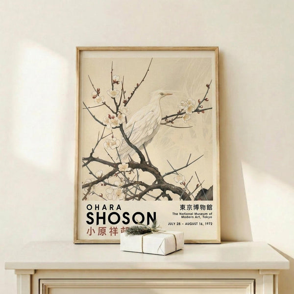May include: Vintage Japanese art poster in light wood frame showing white bird perched on flowering cherry blossom branches against cream background. Exhibition poster features black text for Ohara Shoson at Tokyo National Museum, July-August 1972. Displayed on light wood shelf with small wrapped gift box, against neutral wall with soft natural lighting.