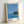May include: Framed art print leaning against white wall showing coastal scene with blue ocean, small rocky island with dark vegetation, clear blue sky, and white waves. Bottom features artist name in black text with colored dots and Tokyo 1973. Natural wood frame with white matting.