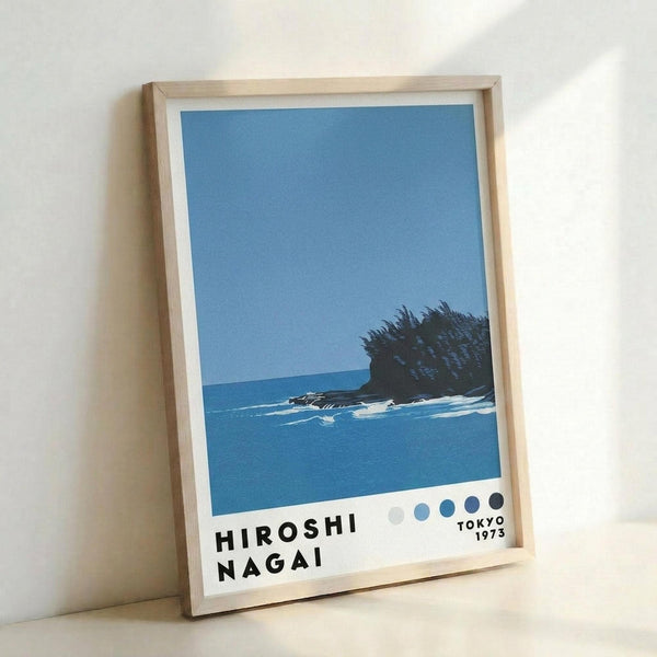 May include: Framed art print leaning against white wall showing coastal scene with blue ocean, small rocky island with dark vegetation, clear blue sky, and white waves. Bottom features artist name in black text with colored dots and Tokyo 1973. Natural wood frame with white matting.