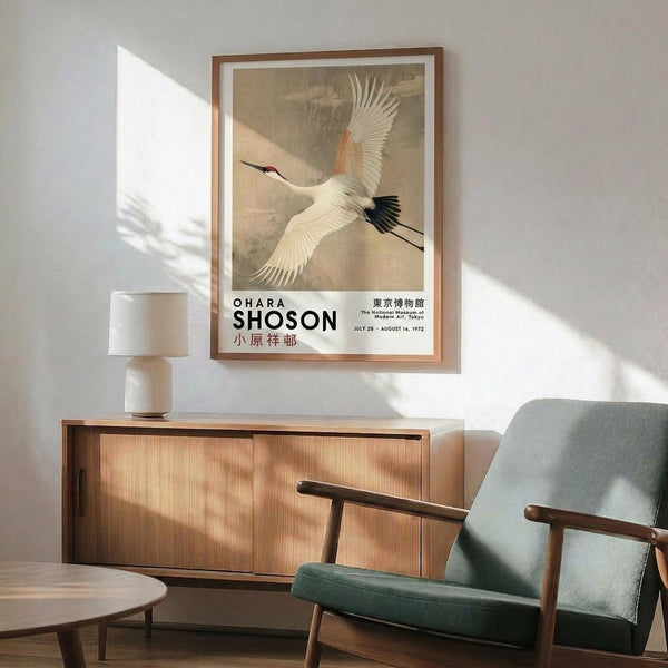 May include: Framed vintage Japanese crane poster hanging on white wall above wooden sideboard with ceramic table lamp. Mid-century modern sage green armchair with wooden arms sits nearby on light wooden floor. Natural sunlight creates diagonal shadows across the minimalist room scene. The poster features a white crane in flight with outstretched wings against a muted beige background in traditional Asian art style.