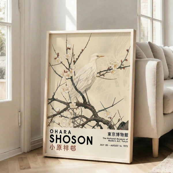 May include: Framed vintage Japanese art poster featuring white bird perched on flowering cherry blossom branches against cream background, displayed on light wood floors next to beige upholstered sofa in bright room with white walls and large windows casting natural light shadows.