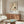 May include: Framed vintage Japanese art poster featuring a white crane with orange and black wing details in flight against a muted beige sky, displayed above a mid-century walnut credenza in a minimalist living room with cream walls, white upholstered chair, brass floor lamp, and oval glass coffee table