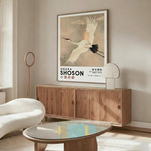 May include: Framed vintage Japanese art poster featuring a white crane with orange and black wing details in flight against a muted beige sky, displayed above a mid-century walnut credenza in a minimalist living room with cream walls, white upholstered chair, brass floor lamp, and oval glass coffee table