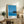 May include: Framed poster with blue coastal scene showing ocean waves and silhouetted tropical vegetation against clear sky, displayed on rustic wooden sideboard alongside potted fern, ceramic vases, and books in warm-toned living room setting with beige upholstered furniture