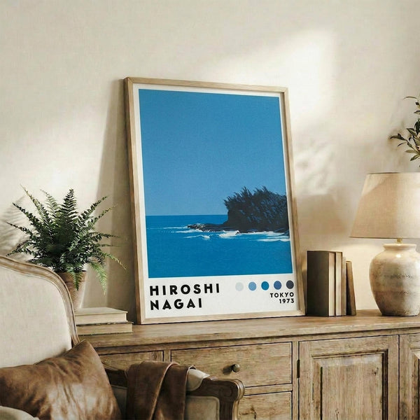 May include: Framed poster with blue coastal scene showing ocean waves and silhouetted tropical vegetation against clear sky, displayed on rustic wooden sideboard alongside potted fern, ceramic vases, and books in warm-toned living room setting with beige upholstered furniture