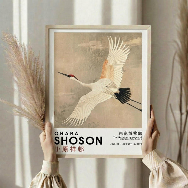 May include: Person in beige clothing holding framed poster featuring Japanese-style painting of white crane with orange neck and black wing tips in flight against cloudy beige background. Dried pampas grass visible on left. Text shows museum exhibition details from Tokyo 1972. Light wooden frame with white mat border.