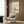 May include: Beige armchair with geometric pillow positioned against wood-paneled wall, displaying framed vintage Japanese art poster featuring white bird perched on flowering branch. Modern floor lamp with brass finish and marble base stands nearby next to dark round side table with open book. Natural lighting from window with sheer curtains creates warm, contemporary living room setting.