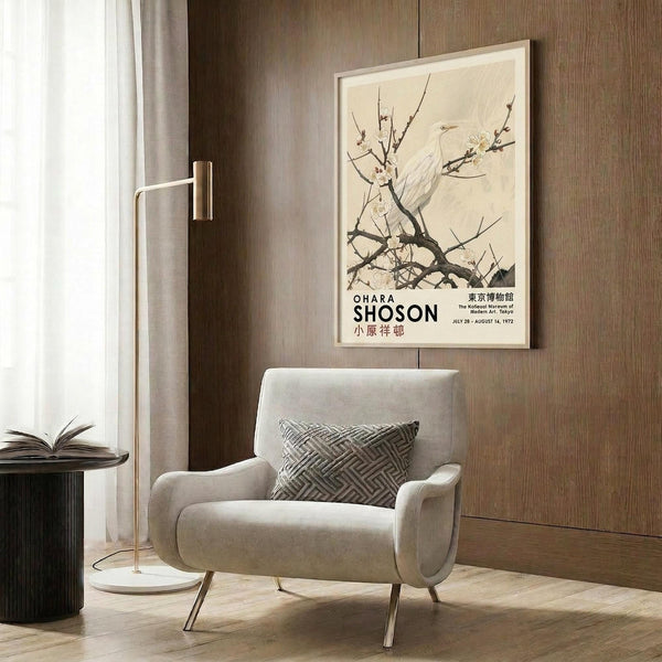 May include: Beige armchair with geometric pillow positioned against wood-paneled wall, displaying framed vintage Japanese art poster featuring white bird perched on flowering branch. Modern floor lamp with brass finish and marble base stands nearby next to dark round side table with open book. Natural lighting from window with sheer curtains creates warm, contemporary living room setting.