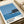 May include: Framed art print showing minimalist blue seascape with dark silhouetted island or rock formation, white wave foam, and clear sky. Print features "HIROSHI NAGAI" text and "TOKYO 1973" with colored dots in corner. Displayed on light wooden surface next to rolled cream paper, clean modern aesthetic.