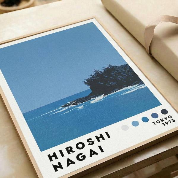 May include: Framed art print showing minimalist blue seascape with dark silhouetted island or rock formation, white wave foam, and clear sky. Print features "HIROSHI NAGAI" text and "TOKYO 1973" with colored dots in corner. Displayed on light wooden surface next to rolled cream paper, clean modern aesthetic.