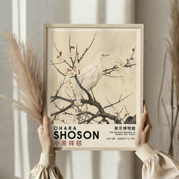 May include: Person holding framed vintage Japanese art poster featuring white bird perched on flowering cherry blossom branch against cream background. Light wood frame, museum exhibition text at bottom. Neutral beige setting with dried pampas grass visible. Hands wearing cream ruffled sleeves display the botanical artwork in soft natural lighting.