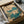 May include: Framed botanical print featuring pink lotus flowers with large teal and green leaves on cream background, displayed in black frame nestled in cardboard shipping box with white tissue paper and wooden rolling tubes visible alongside