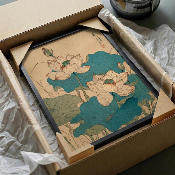 May include: Framed botanical print featuring pink lotus flowers with large teal and green leaves on cream background, displayed in black frame nestled in cardboard shipping box with white tissue paper and wooden rolling tubes visible alongside