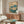 May include: Japanese botanical artwork featuring white lotus flowers with large teal leaves displayed above a mid-century walnut credenza in a minimalist living room. The vintage-style print shows delicate blooms and buds against a cream background, framed in thin wood. Modern white curved chair and glass coffee table complete the serene, natural-toned interior setting.