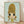 May include: Framed art print displayed on wooden floor against white wall with natural light shadows. Shows cartoon-style character with oversized golden blonde beehive hairstyle, large dark eyes, and yellow top against soft mint green background. Artwork has retro aesthetic with muted color palette in light wood frame.