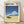 May include: Framed poster print leaning against white wall on wooden floor. Features retro-style artwork with blue sky, white puffy cloud, ocean waves, and dark vintage car on sandy beach. Japanese text at top. Light wood frame with cream matting. Natural lighting creates soft shadows on textured white wall.