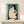 May include: Framed Japanese woodblock print featuring a nude woman with black hair combing her dark hair against a textured blue-green background, displayed on a light wood shelf beside a small wrapped gift with natural twine and dried grass accent, set against a cream colored wall.