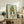 May include: Framed art print featuring anime-style character with golden blonde beehive hairstyle and large dark eyes on mint green background, displayed on rustic wooden sideboard alongside fern plant, books, and ceramic pottery in neutral-toned living room setting with beige sofa and white walls.