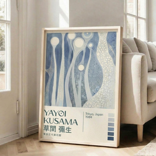May include: Framed exhibition poster featuring abstract blue and white organic shapes with dotted patterns, leaning against white wall next to beige upholstered chair. Poster displays Japanese text and "Tokyo, Japan 1984" with blue color swatches at bottom. Natural sunlight streams through windows creating shadows on wooden floor.