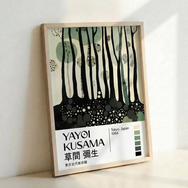 May include: Framed poster print leaning against white wall showing abstract forest scene with vertical black tree trunks, sage green circular shapes, dotted patterns, and speckled ground in black and white. Bottom features exhibition text "YAYOI KUSAMA" with Japanese characters, "Tokyo, Japan 1984" and color palette strip. Natural wood frame, soft lighting creates gentle shadows on wall.
